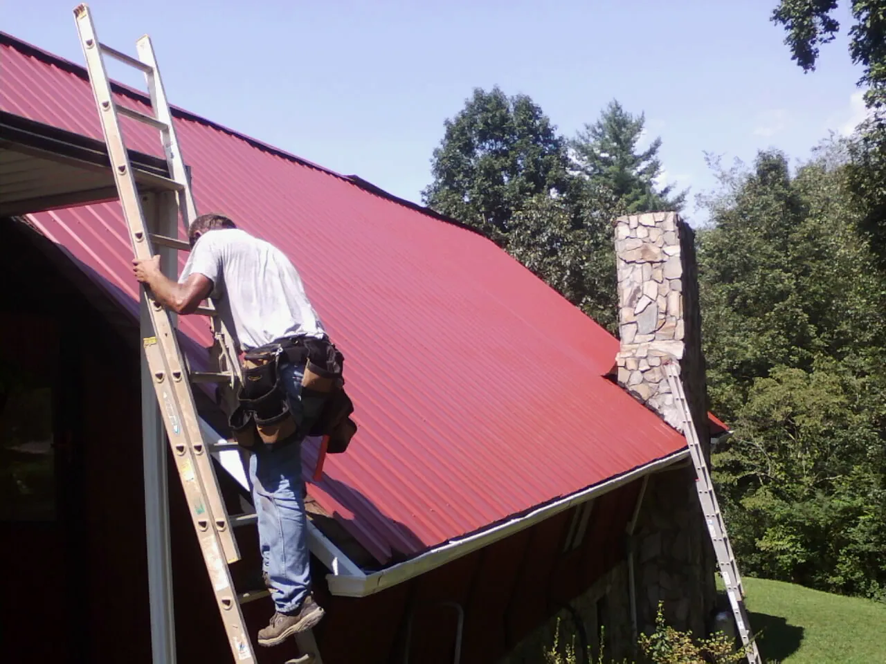 Roofer on a ladder beside a red metal roof and stone chimney — Soffit & Fascia Repair by Roofing Brothers in Dekalb
